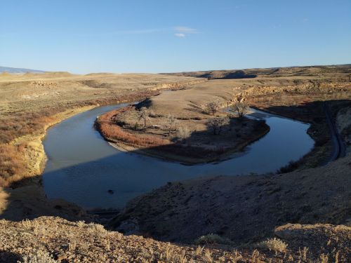 A winding river flows through a dry, hilly landscape under a clear blue sky. Sparse vegetation lines the banks.