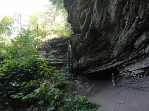 A person stands near a rocky overhang with a small waterfall surrounded by lush greenery.