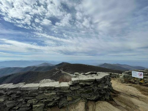 A panoramic view from a rocky mountain summit, featuring rolling hills and a cloudy sky. A stone wall is in the foreground.