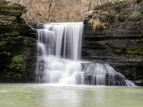 A serene waterfall cascades over rocky cliffs into a calm, green pool surrounded by autumn foliage.