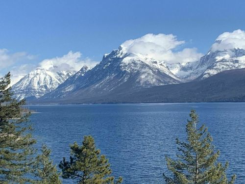 Snow-capped mountains rise above a serene blue lake, framed by green trees under a clear blue sky.