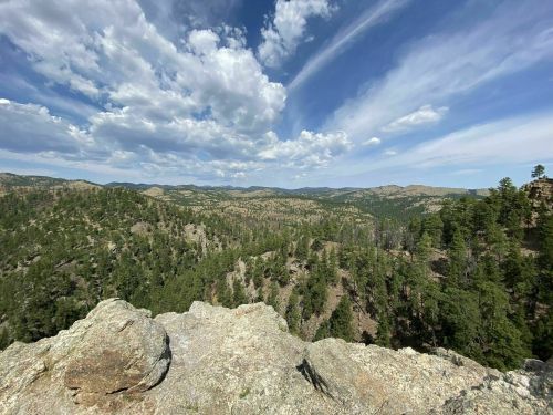 A panoramic view of lush green mountains under a blue sky with scattered clouds. Rocky foreground adds depth to the scene.
