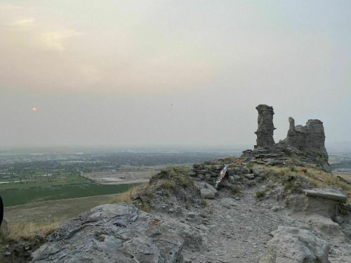 A rocky outcrop with two tall formations overlooks a hazy landscape at sunset, with fields stretching into the distance.