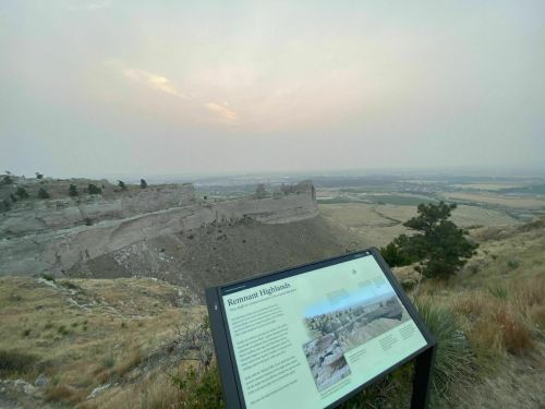 A scenic view of a rocky cliff with a sign about the Remnant Highlands, surrounded by hazy skies and distant fields.