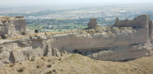 A panoramic view of a rocky cliff overlooking a valley and a small town in the distance, under a hazy sky.