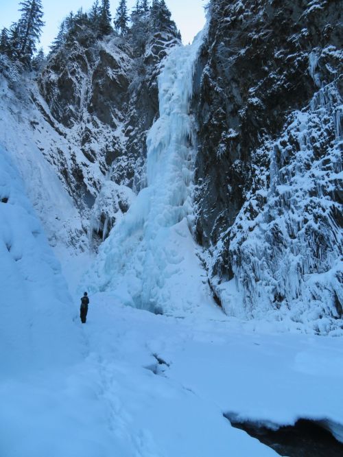 A person stands near a frozen waterfall surrounded by snow-covered cliffs and trees.