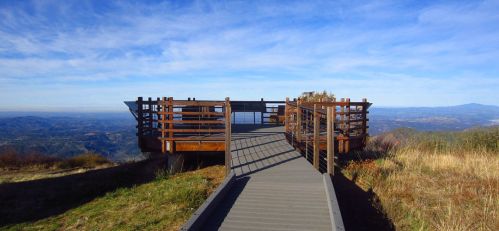 A wooden observation deck on a hillside, overlooking a vast landscape under a clear blue sky.
