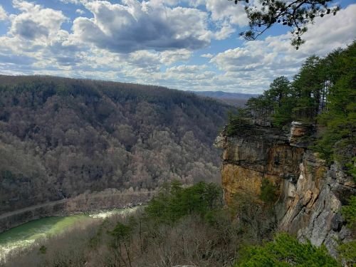 A scenic view of a rocky cliff overlooking a winding river, surrounded by trees and rolling hills under a cloudy sky.