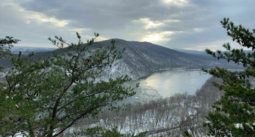 A scenic view of a river winding through snow-covered hills under a cloudy sky, framed by evergreen trees.