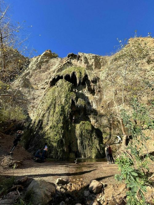 A lush waterfall cascades down a rocky cliff, with people exploring the area and enjoying the natural scenery.