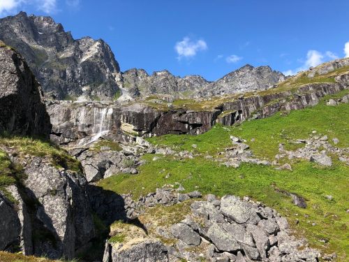A scenic mountain landscape featuring rocky cliffs, green grass, and a waterfall under a clear blue sky.
