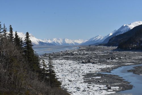 A scenic view of a riverbank with ice, mountains, and clear blue skies in a remote, natural landscape.