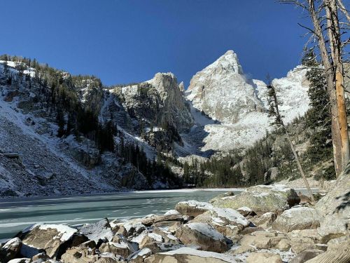 Snow-covered mountains rise above a frozen lake, surrounded by rocky terrain and evergreen trees under a clear blue sky.