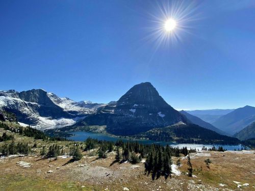 A stunning mountain landscape with a clear blue sky, sun shining, and a lake surrounded by trees and rocky terrain.