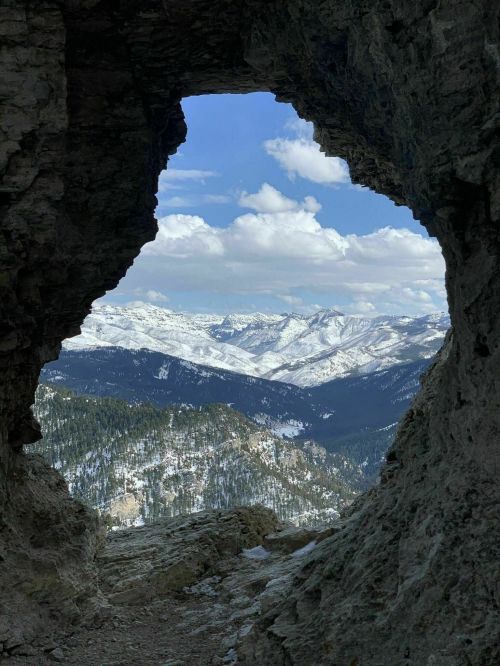 View through a rocky archway, showcasing snow-capped mountains and a blue sky with scattered clouds.