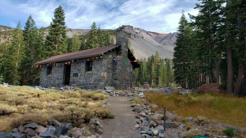 A stone cabin surrounded by tall trees and mountains, with colorful prayer flags hanging in the breeze.