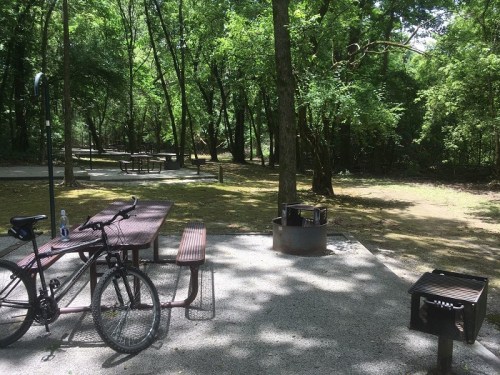 A shaded picnic area with a bike, tables, and a grill surrounded by trees in a peaceful park setting.