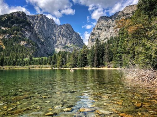 A serene lake surrounded by tall mountains and lush trees under a blue sky with fluffy clouds.