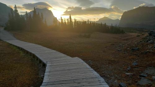 A wooden pathway leads through a serene landscape at sunset, surrounded by mountains and trees.