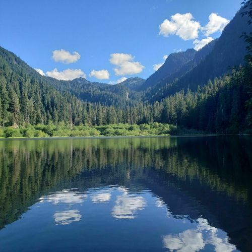 A serene lake surrounded by lush green mountains and blue skies, reflecting clouds on the water's surface.