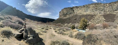 A person stands on a rock in a canyon, surrounded by dry vegetation and cliffs under a partly cloudy sky.