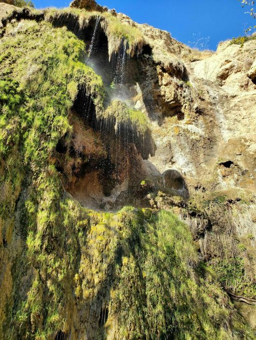 A cascading waterfall flows down a rocky cliff covered in green moss under a clear blue sky.