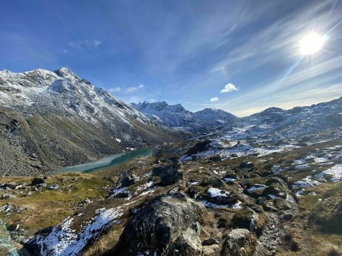 A scenic mountain landscape with snow-capped peaks, a turquoise lake, and a bright sun in a clear blue sky.