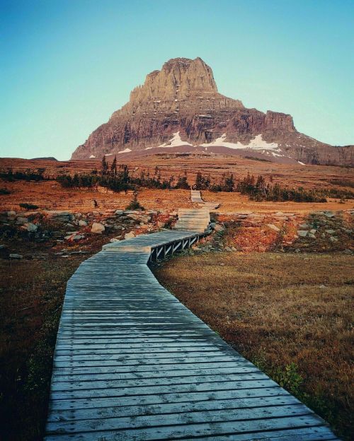 A wooden boardwalk leads through a grassy landscape towards a towering mountain under a clear blue sky.