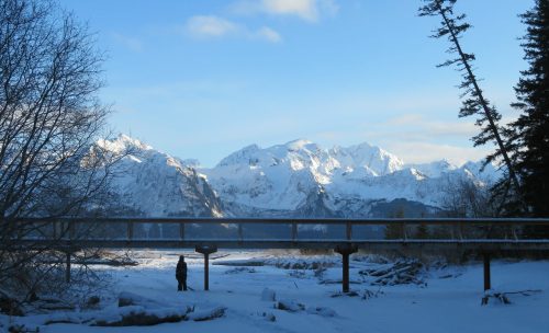 A person stands on a snowy landscape beneath a clear blue sky, with snow-capped mountains in the background.
