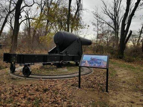 A historic cannon on display outdoors, with a nearby informational sign and trees in the background.