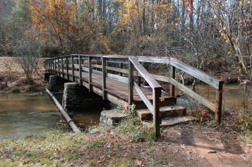 A wooden bridge spans a calm stream, surrounded by autumn foliage and trees.