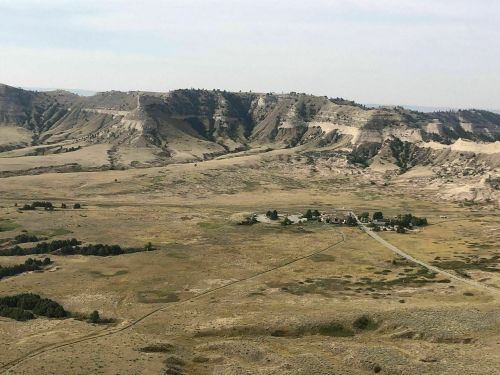A vast, dry landscape with rolling hills and a small cluster of buildings in the foreground.