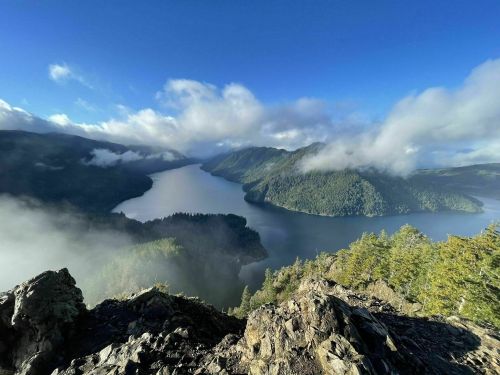 A panoramic view of a serene lake surrounded by lush mountains and misty clouds under a clear blue sky.