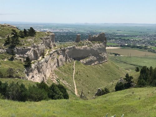 A scenic view of a rocky cliff with greenery, overlooking a valley and distant hills under a clear blue sky.