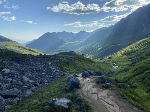 A scenic mountain landscape with a winding path, rocky terrain, and lush green hills under a blue sky with clouds.