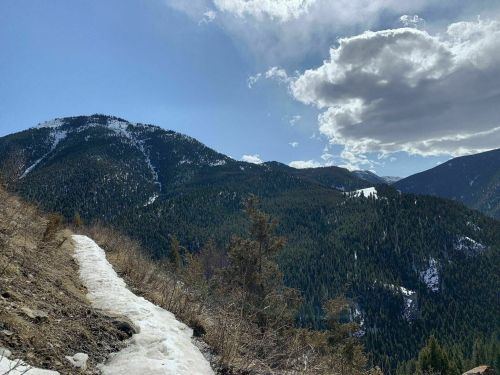 A snow-covered trail winds through a mountainous landscape with lush green trees and a blue sky dotted with clouds.