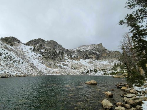 A serene mountain landscape with a snowy peak reflected in a calm lake, surrounded by rocky shores and trees.