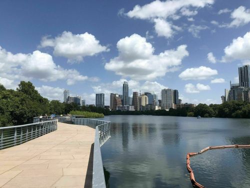A scenic view of a city skyline reflected in a calm lake, with a walkway and fluffy clouds in a blue sky.