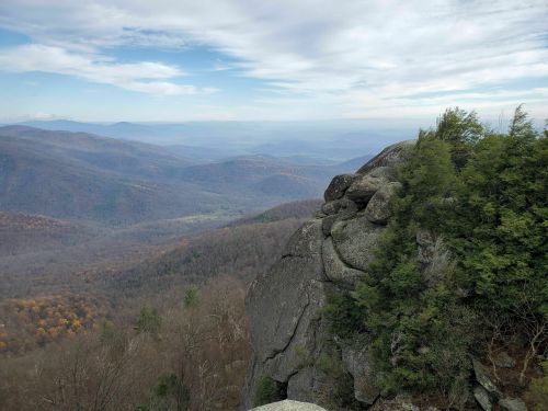 A rocky outcrop overlooks a vast valley with rolling hills and distant mountains under a cloudy sky.