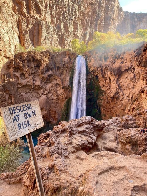 A waterfall cascades down rocky cliffs, with a wooden sign warning "Descend at Own Risk" nearby.