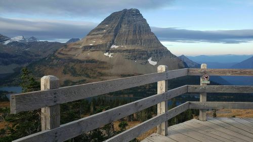 A wooden viewing platform overlooks a mountain peak with a rugged, rocky face and a clear blue sky in the background.