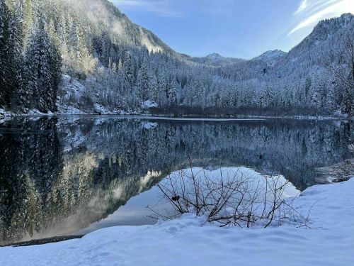 A serene winter landscape featuring a snow-covered lake surrounded by mountains and evergreen trees, reflecting the scenery.