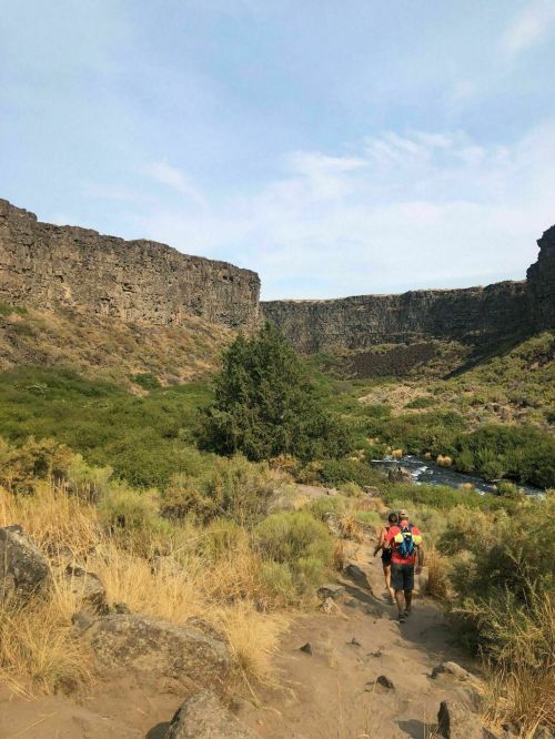 A hiker walks along a sandy path through a canyon, with rocky cliffs and a river visible in the background.