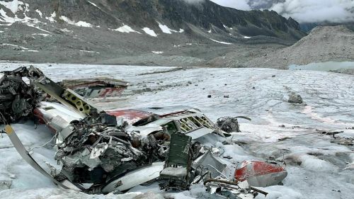 Wreckage of an aircraft on a glacier, surrounded by rocky terrain and snow-capped mountains.