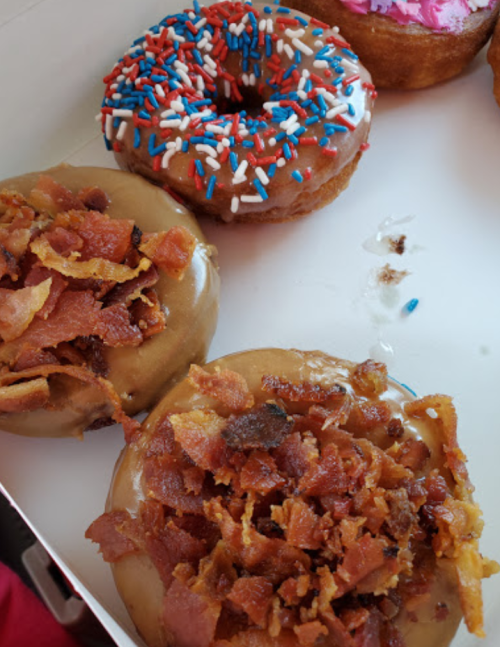 A box of donuts featuring a glazed donut topped with bacon and a red, white, and blue sprinkled donut.