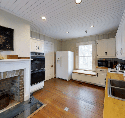 Bright kitchen with white cabinets, a fireplace, and a window seat, featuring modern appliances and wooden flooring.