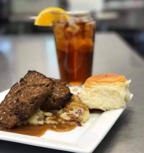 A plate of meatloaf with gravy, mashed potatoes, a biscuit, and a glass of iced tea with a slice of orange.