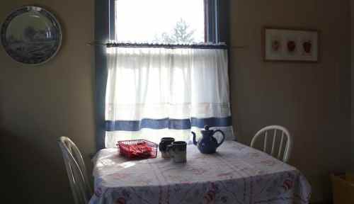 A cozy kitchen scene with a table covered in a floral tablecloth, a teapot, and a window with sheer curtains.