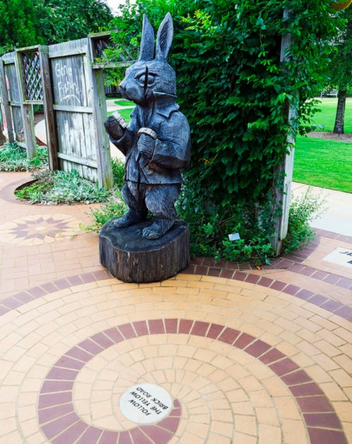 A wooden rabbit statue stands on a circular brick path, surrounded by greenery and a wooden fence.