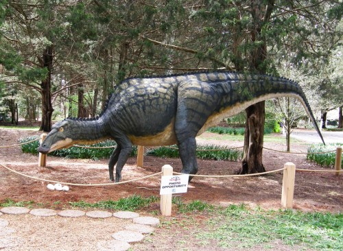 A life-sized dinosaur sculpture stands in a wooded area, surrounded by greenery and a sign for photo opportunities.